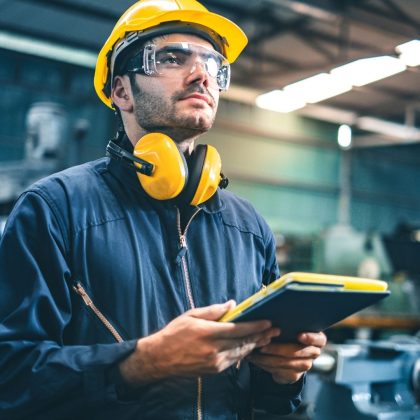 Man in a construction environment wearing safety glasses, hearing protection and holding a clipboard whilst wearing overalls